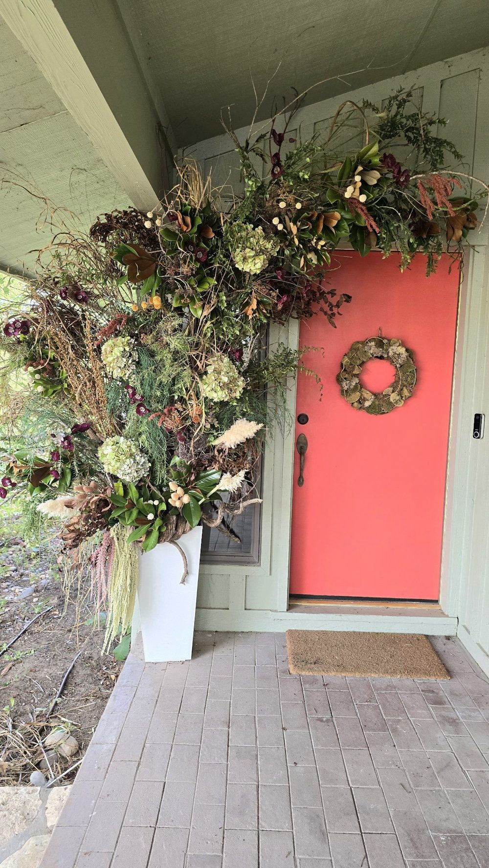 Completed botanical arch installation framing a red front door with dried botanicals, grasses, and a foraged wreath